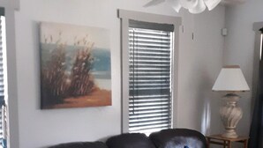 Een woonkamer met een donker leren bank, een plafondventilator, een raam met jaloezieën, een schilderij aan de muur en een lamp op een bijzettafeltje.