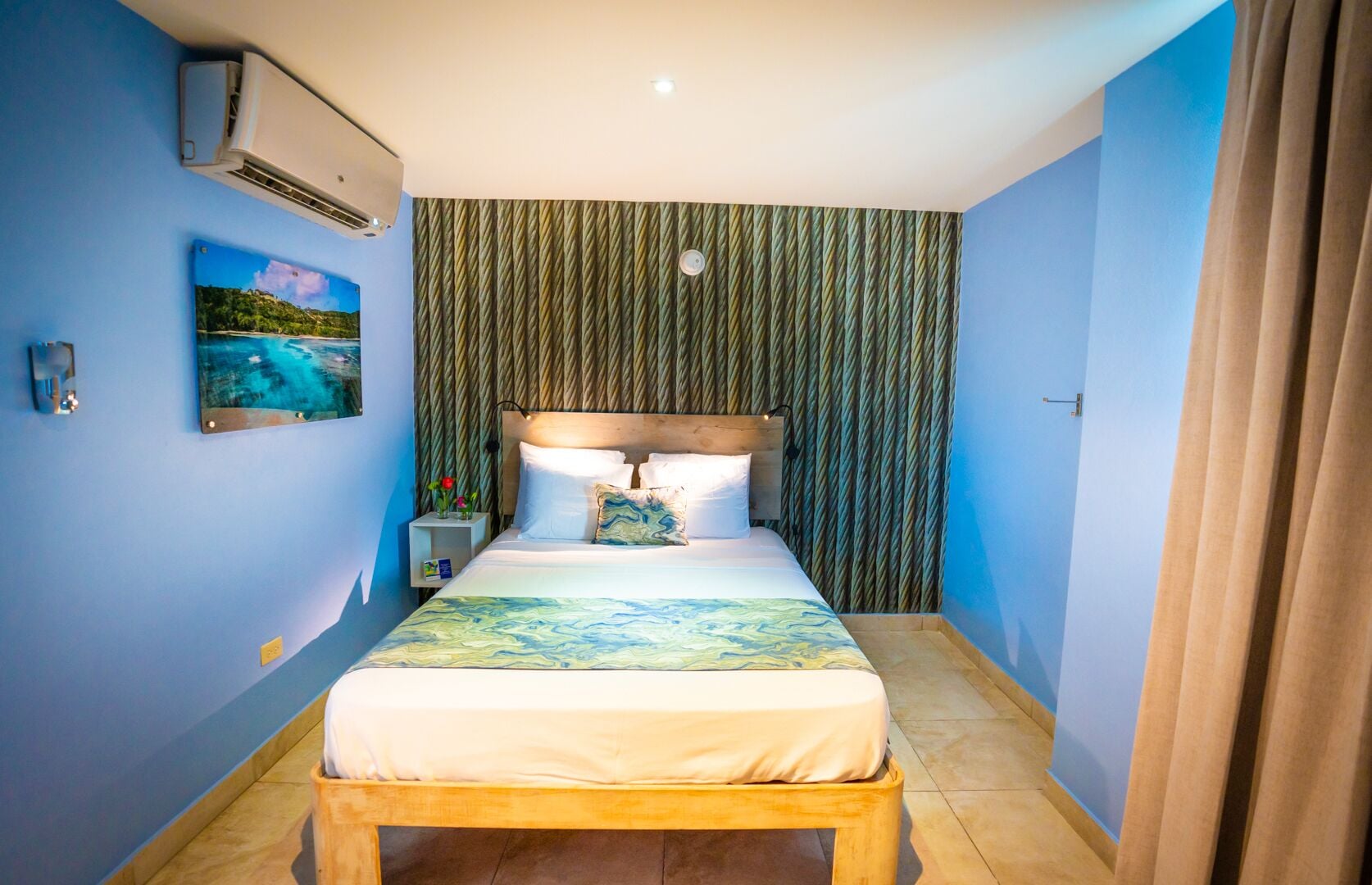 A bedroom with a bed, a nightstand, a wall-mounted air conditioner, a painting, and a patterned curtain.