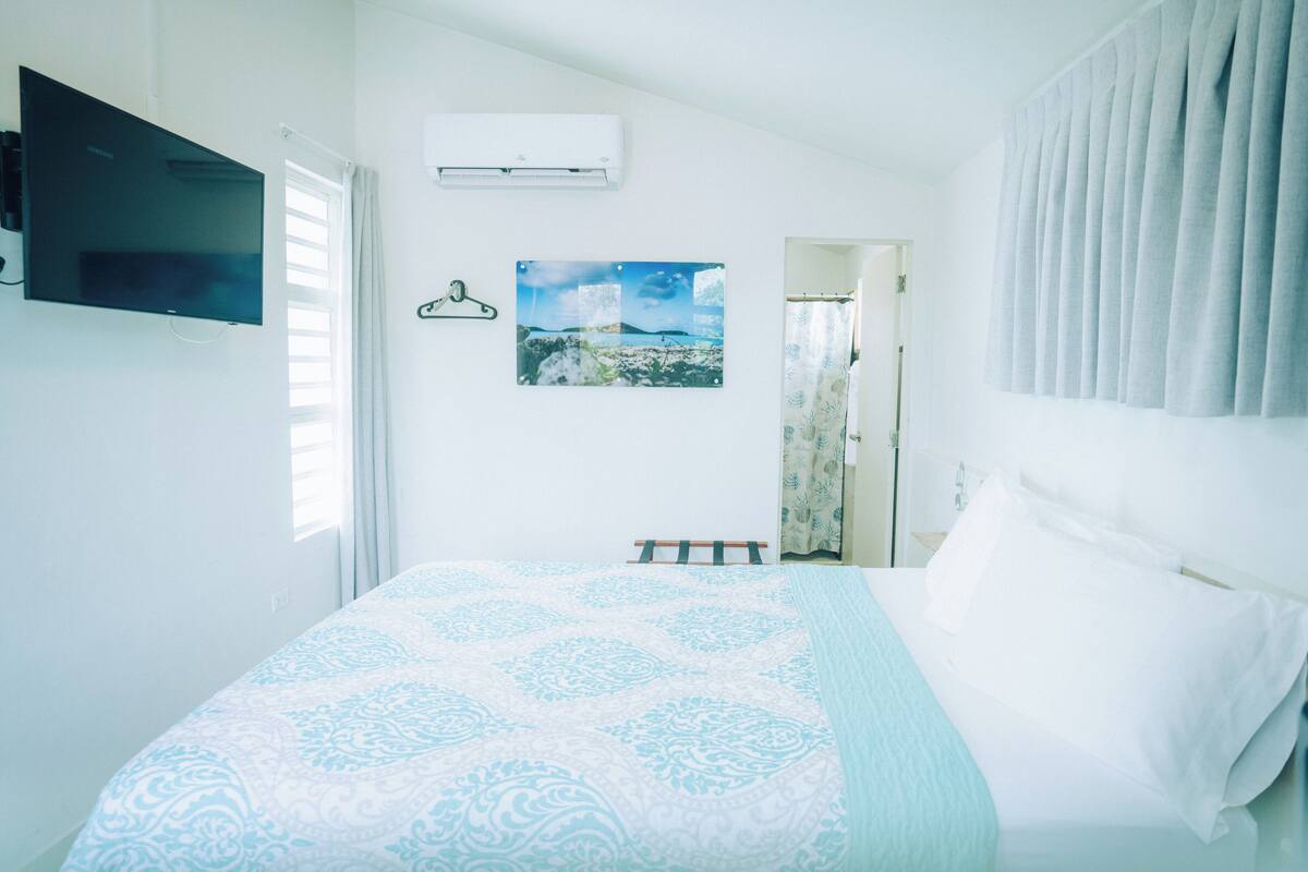 Een slaapkamer met een bed, een aan de muur bevestigde televisie, een airconditioner en uitzicht op buiten door een raam.