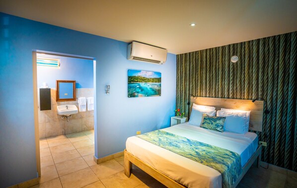 A hotel room with a bed, a nightstand, a wall-mounted air conditioner, a bathroom with a sink and towels, and a painting of a beach scene.