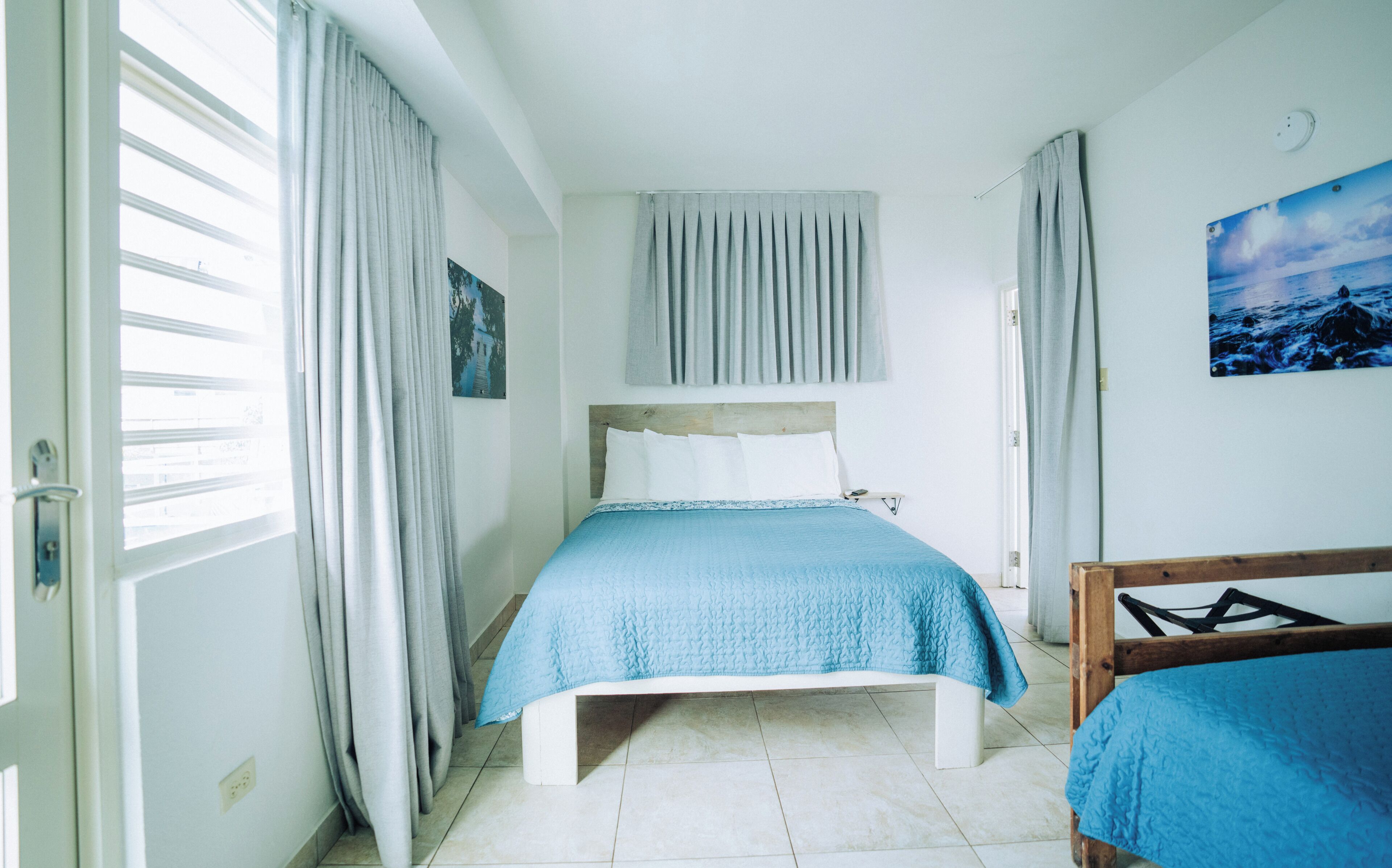 A bedroom with a bed, a nightstand, a window with curtains, a wall-mounted light, and a painting.