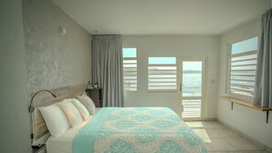 Un dormitorio con cama, mesita de noche, lámpara, ventana con vista al mar y puerta que da a un balcón exterior.