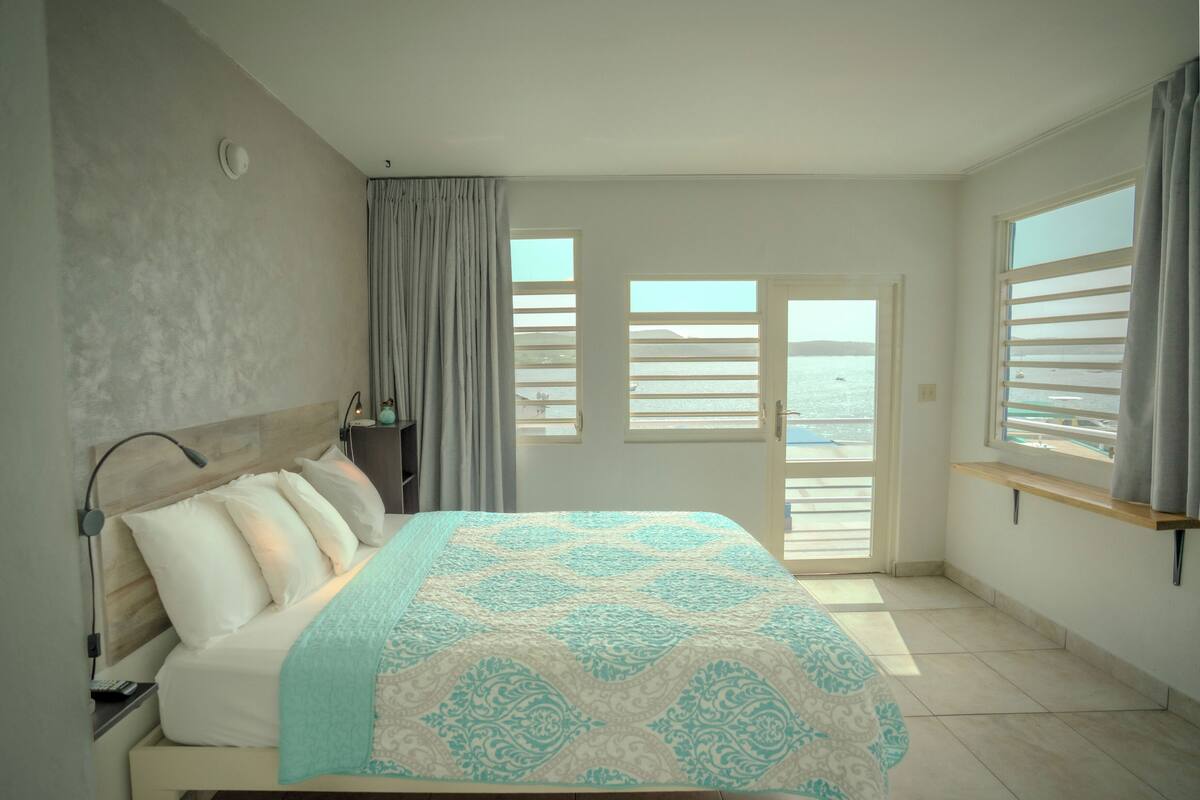 Een slaapkamer met een bed, nachtkastje, lamp, raam met uitzicht op zee en een deur naar een buitenbalkon.