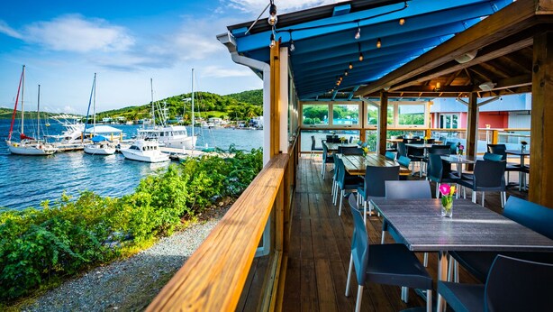 Un área de comedor al aire libre techada con mesas y sillas de madera, con vista a un puerto deportivo lleno de veleros.
