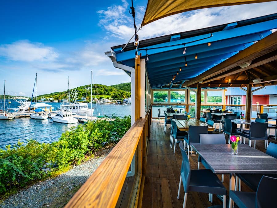 A covered outdoor dining area with wooden tables and chairs, overlooking a marina with sailboats.