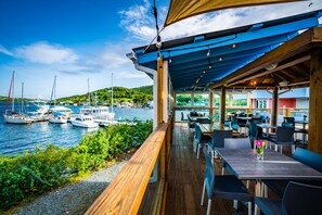 A covered outdoor dining area with wooden tables and chairs, overlooking a marina with sailboats.