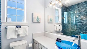 A modern bathroom with a white toilet, a white sink with a blue basin, and a large mirror. There are decorative towels and artwork on the walls. - Wave Whisperer  4br/4ba Steps from the Beach - Pool (Santa Rosa Beach)