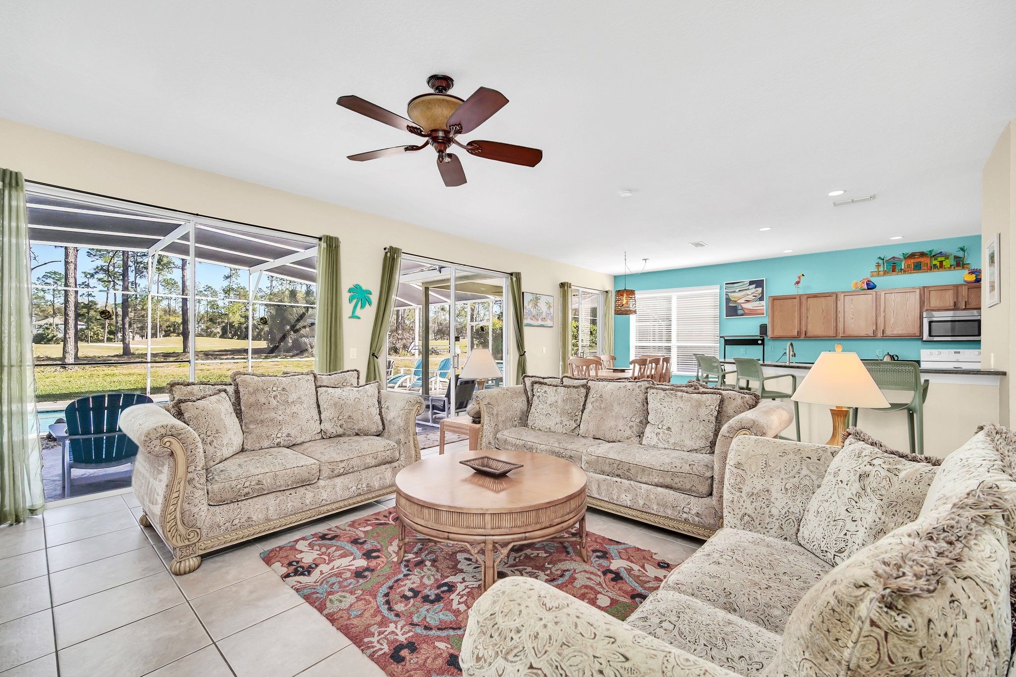 A spacious living room with a large ceiling fan, a round coffee table, and a view of the outdoors through large windows.
