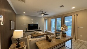 A modern living room with a flat-screen TV, a ceiling fan, and a large window with curtains.