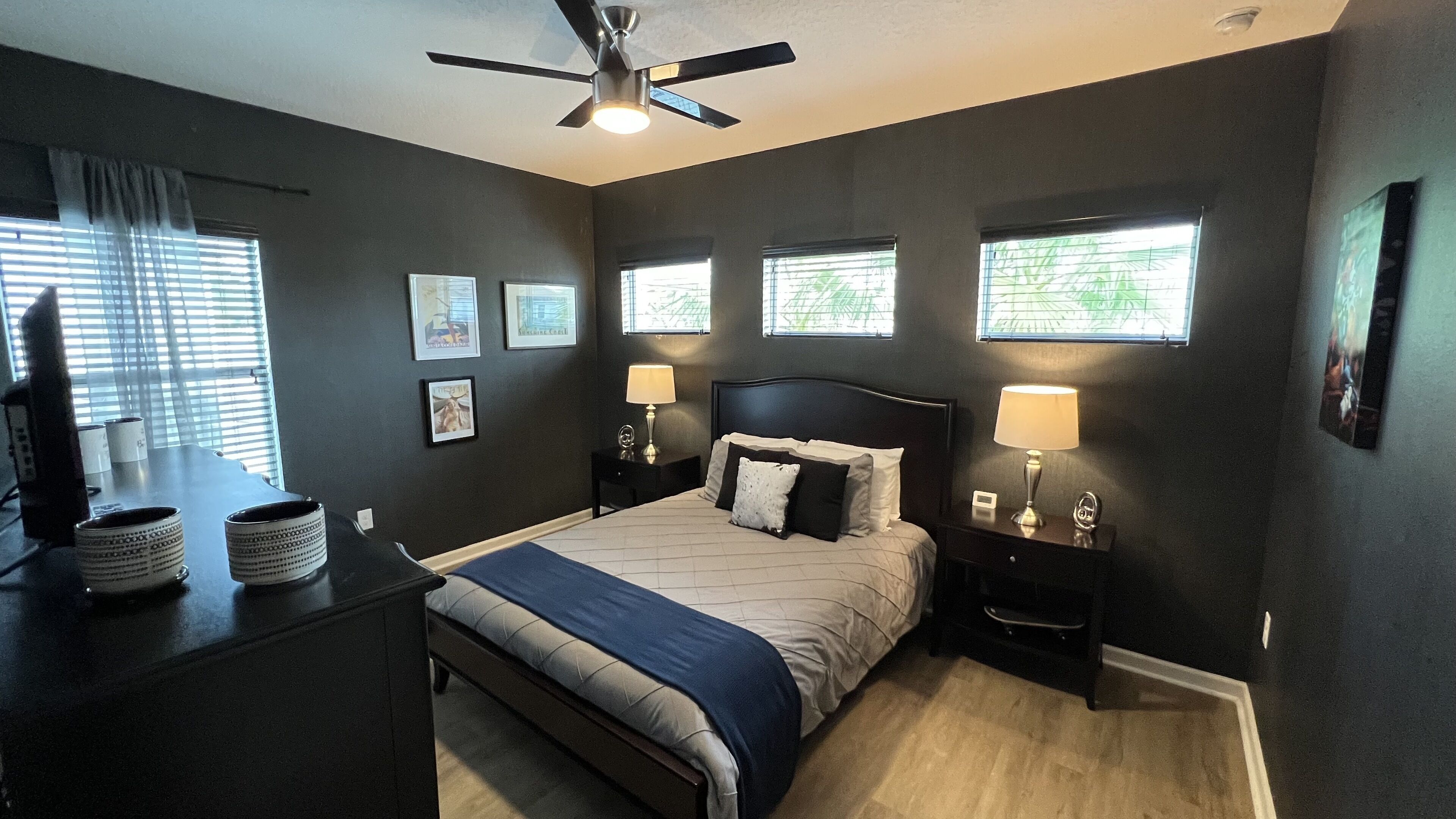 A bedroom with a bed, two bedside tables with lamps, a ceiling fan, and framed pictures on the wall.