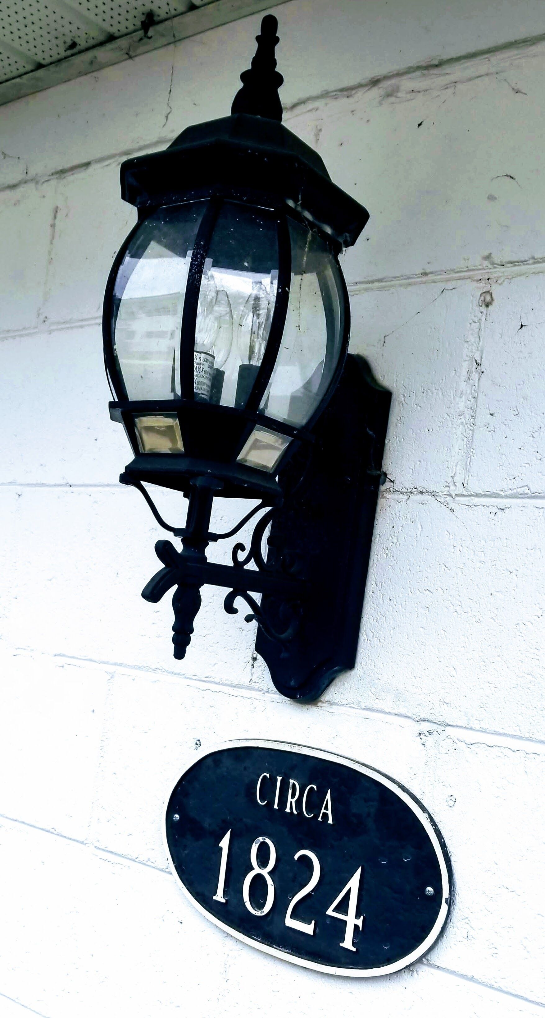Un lampadaire noir, doté d’un abat-jour en verre et d’un socle en métal, est fixé sur un mur blanc. En dessous, une plaque noire et blanche indiquant le numéro de la maison porte l’inscription « VERS 1824 ».