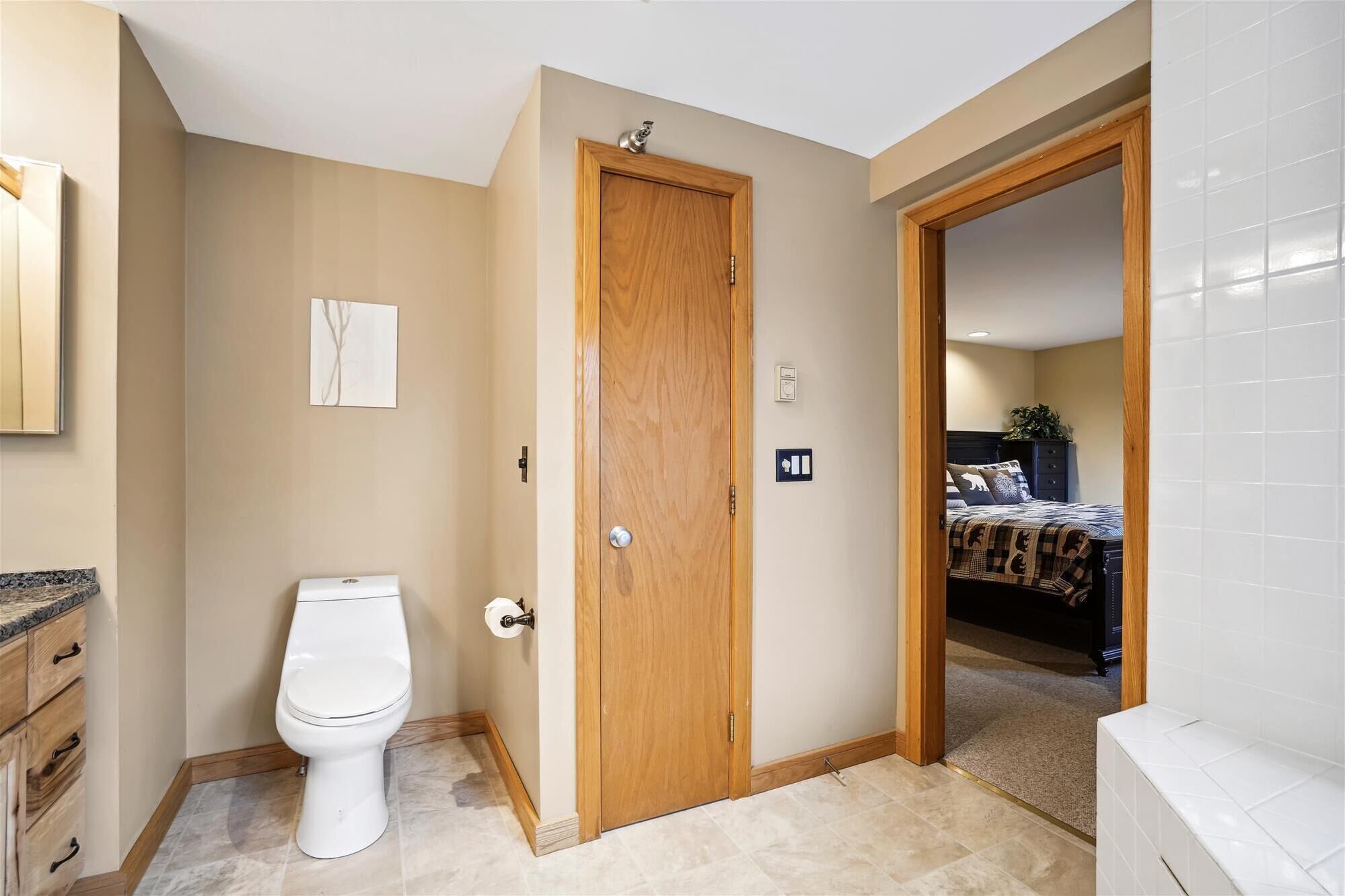 A bathroom with a toilet, wooden door, and a visible bed in the adjacent room.