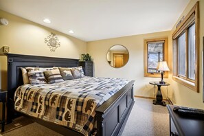 A bedroom with a large bed, a round mirror, a bedside table with a lamp, and a window with blinds. - 2-bedroom plus loft condo with mountain views, pool, and hot tub. (Killington)