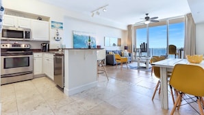 A modern kitchen with white cabinets and stainless steel appliances, a dining area with yellow chairs, and a large window overlooking the ocean. - Gulf Front PH •Free Chairs• Best Views in PCB (Panama City Beach)