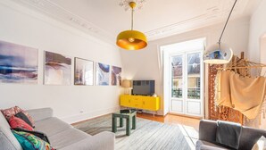 A living room with a grey sofa, a yellow cabinet, and a television. There are abstract paintings on the wall and a window with a view of a balcony. - GuestReady - Groovy Fanqueiros (Lisbon)