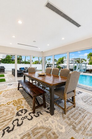 A dining area with a wooden table and chairs, a view of a pool, and a large window. - Luxurious, Modern Fort Lauderdale House with Waterfront Pool & Spa (Fort Lauderdale)