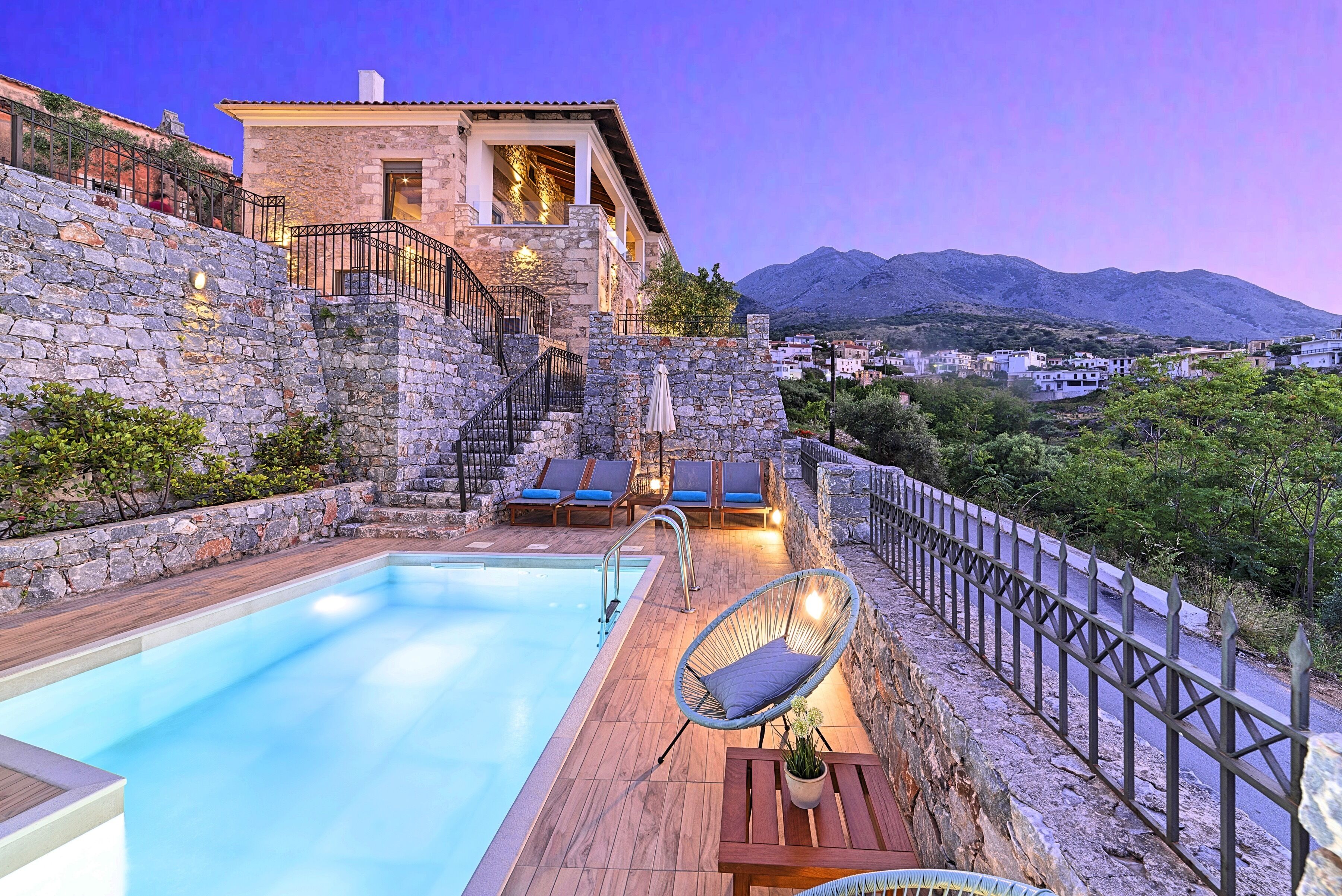 A stone-walled pool area with a wooden deck, lounge chairs, and a view of a mountain range.