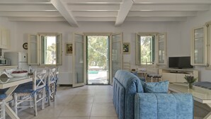 A spacious living room with a sofa, a dining table with chairs, a television, and large windows.