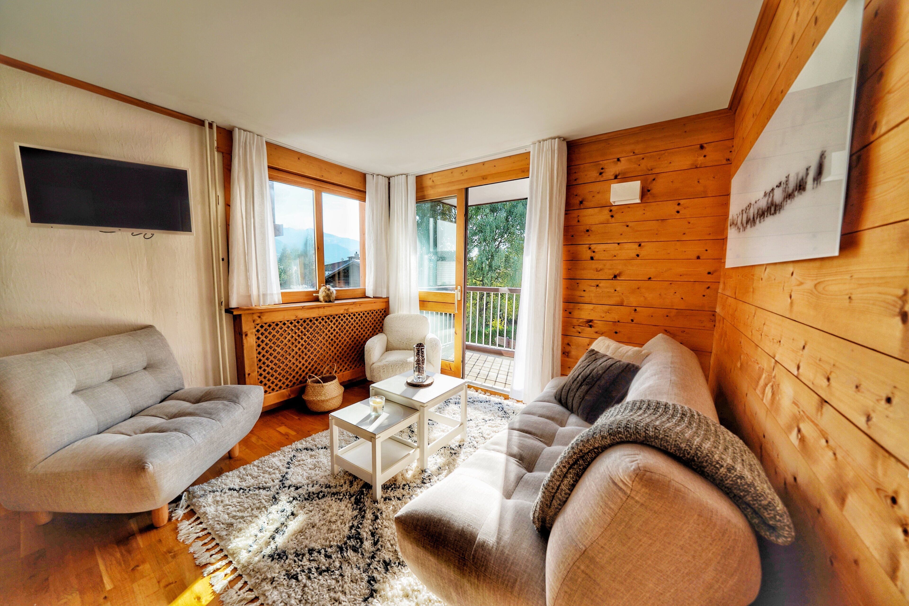 A modern living room with a sofa, armchair, and coffee table. There is a TV mounted on the wall and a window with curtains.