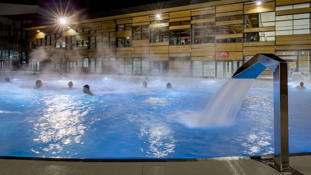 A heated indoor swimming pool with people swimming, a modern building with large windows in the background, and a waterfall feature. - La Ménandière - 3 bedrooms (Megève)