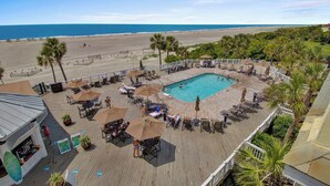 Zona de piscina con sillas de descanso y sombrillas, rodeada por una terraza de madera, con vista a una playa y el océano.