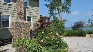 A house with a stone chimney, a gravel driveway, and a garden with pink flowers and green shrubs. - Enjoy the View From this Charming Lakefront House in Private Park (Huron)