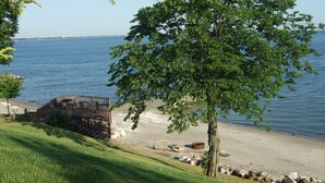 A beach with a wooden deck overlooking the water, a tree in the foreground, and a clear sky. - Enjoy the View From this Charming Lakefront House in Private Park (Huron)