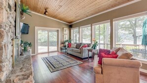 A spacious living room with a stone fireplace, a wooden ceiling, and large windows offering a view of the outdoors.