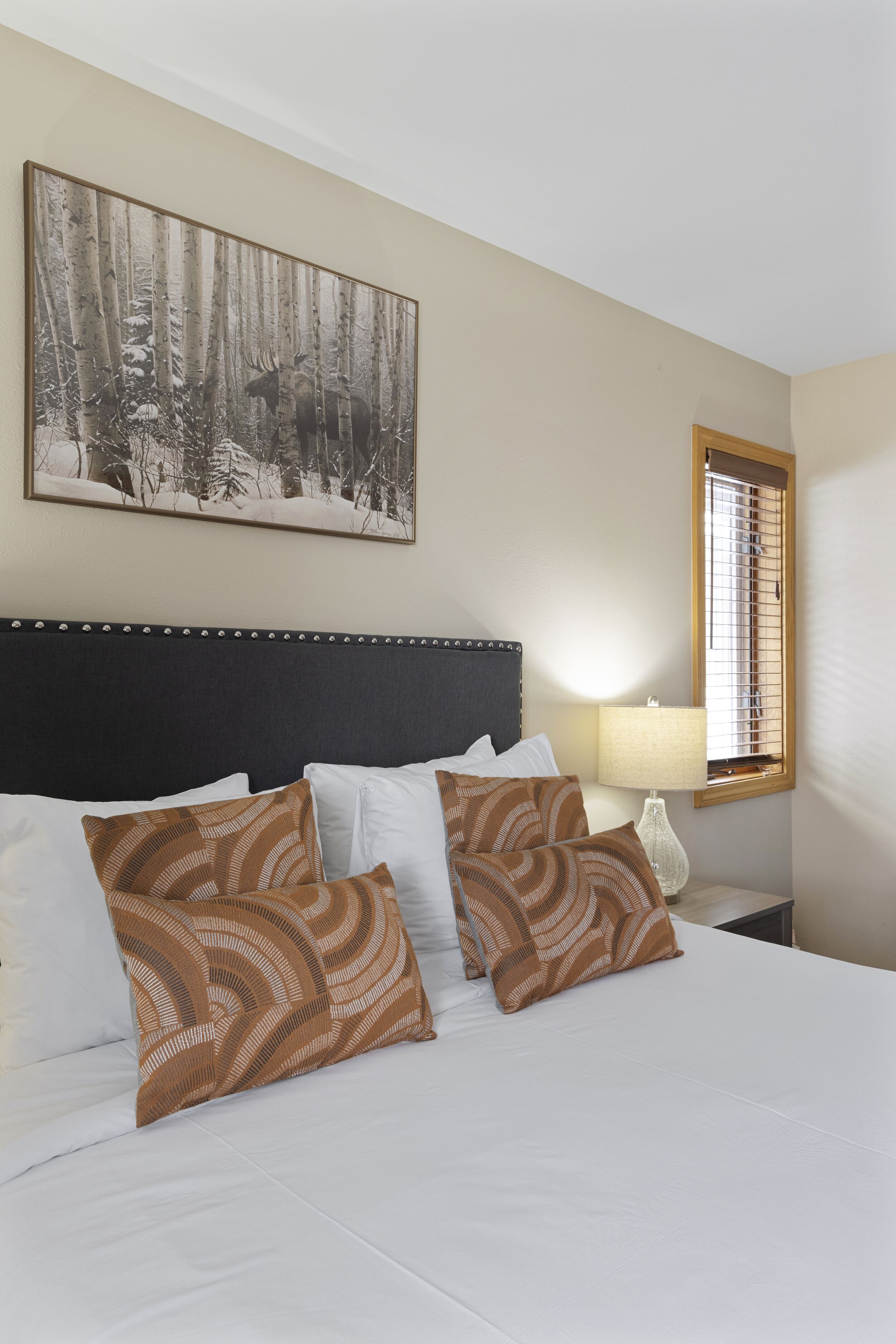 A hotel room with a large bed, a headboard, a bedside table with a lamp, a window with blinds, and a forest-themed painting on the wall.