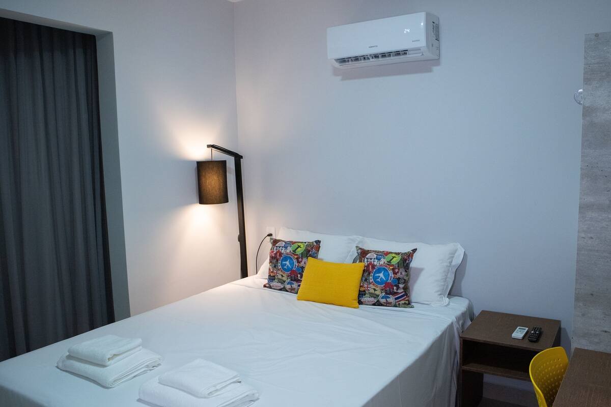 Hotellihuone, jossa on sänky, yöpöytä, lamppu, ilmastointilaite ja pieni kasvi.