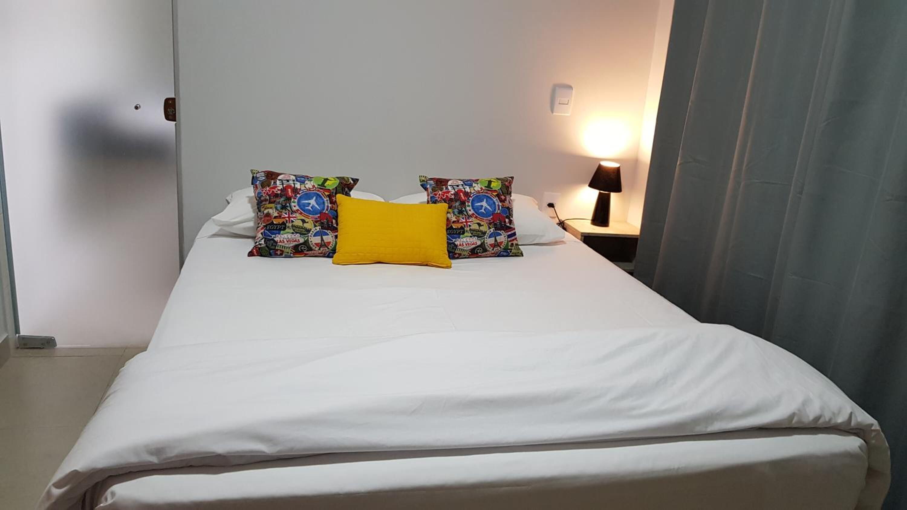 Un lit bien fait, avec du linge de lit blanc, deux coussins décoratifs à motifs évoquant le voyage, et un seul coussin jaune. Une lampe de chevet est allumée sur la droite, et un rideau recouvre partiellement une fenêtre.