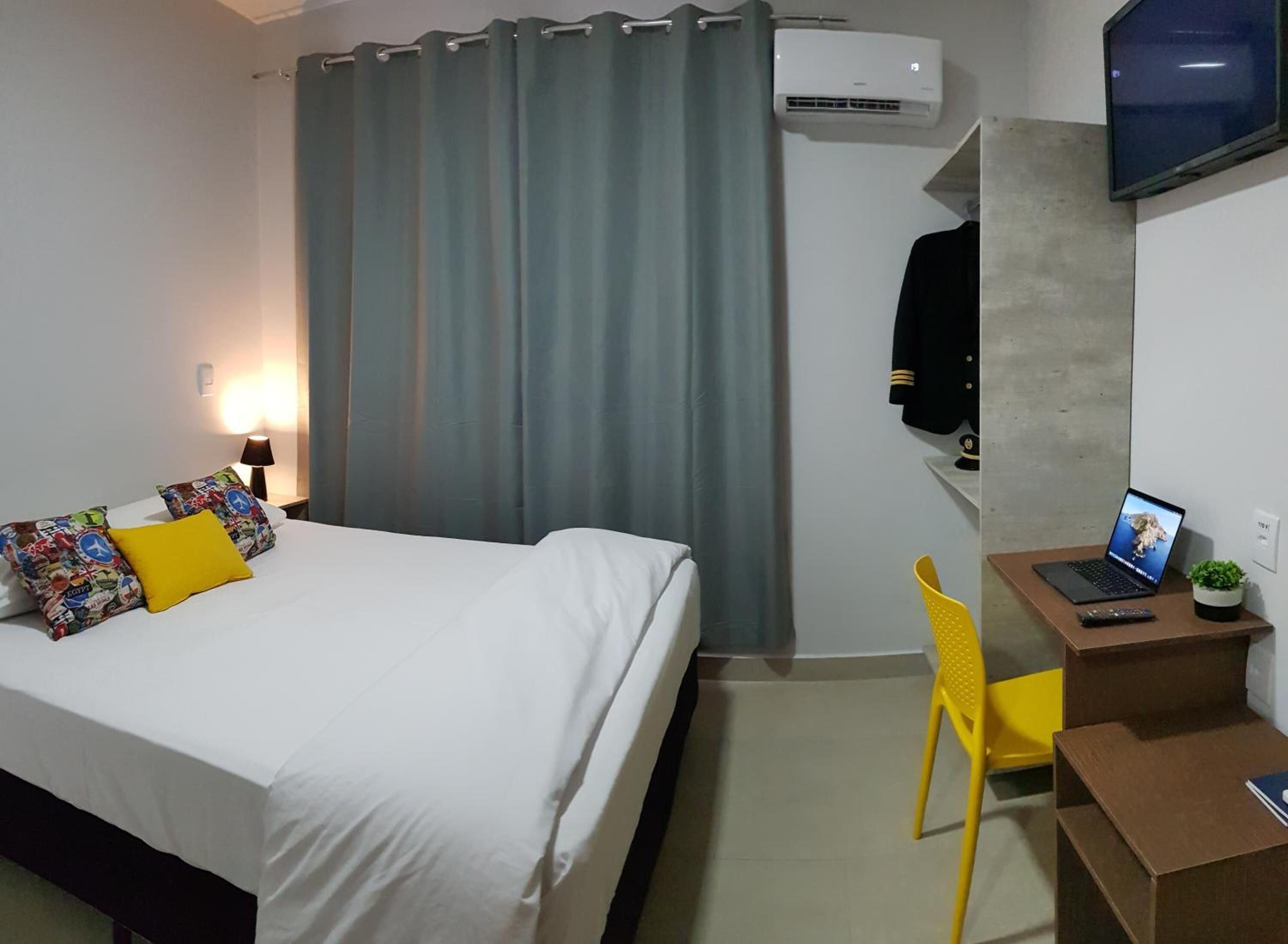 Una habitación de hotel con una cama, un escritorio con una computadora portátil, una silla y un equipo de aire acondicionado.