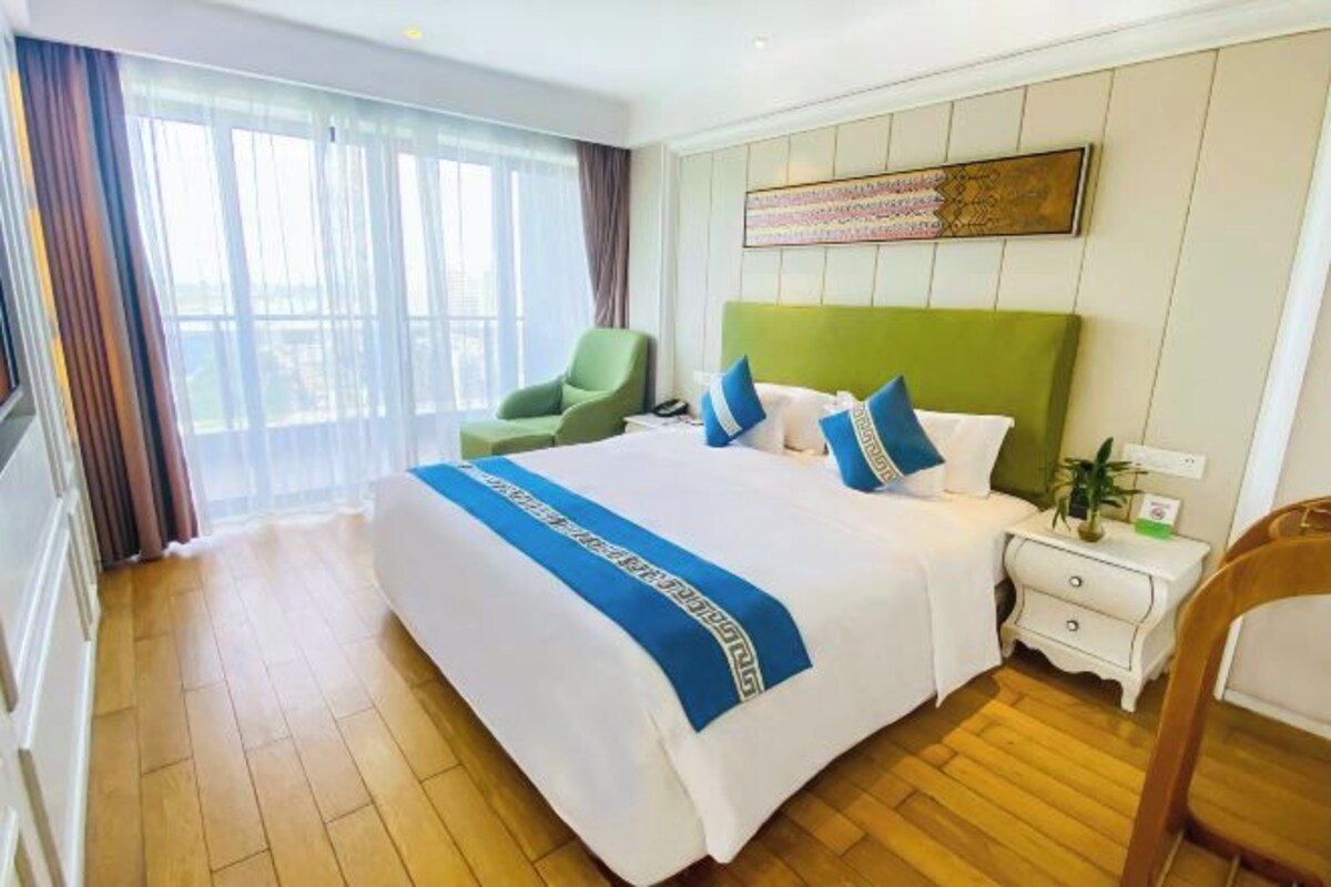Une chambre d’hôtel avec un grand lit, un fauteuil vert, une table de chevet et une fenêtre avec des rideaux.