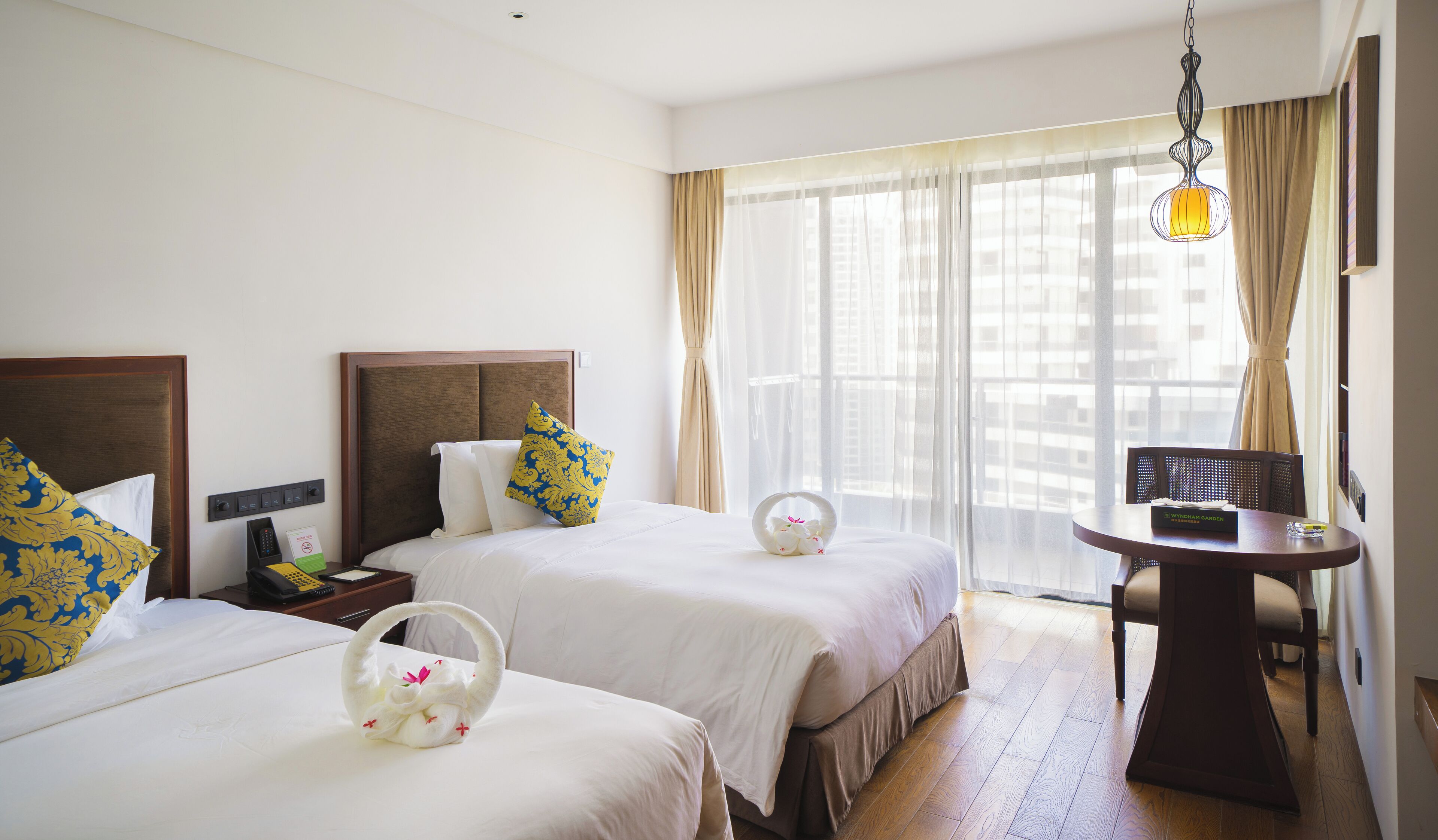 Une chambre d’hôtel avec deux lits, une petite table, une chaise et une fenêtre avec des rideaux légers.