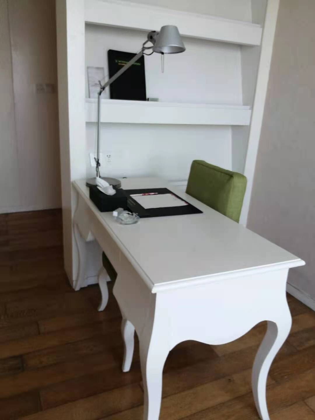A white desk with a lamp, a chair, and a shelf with a book and a phone.
