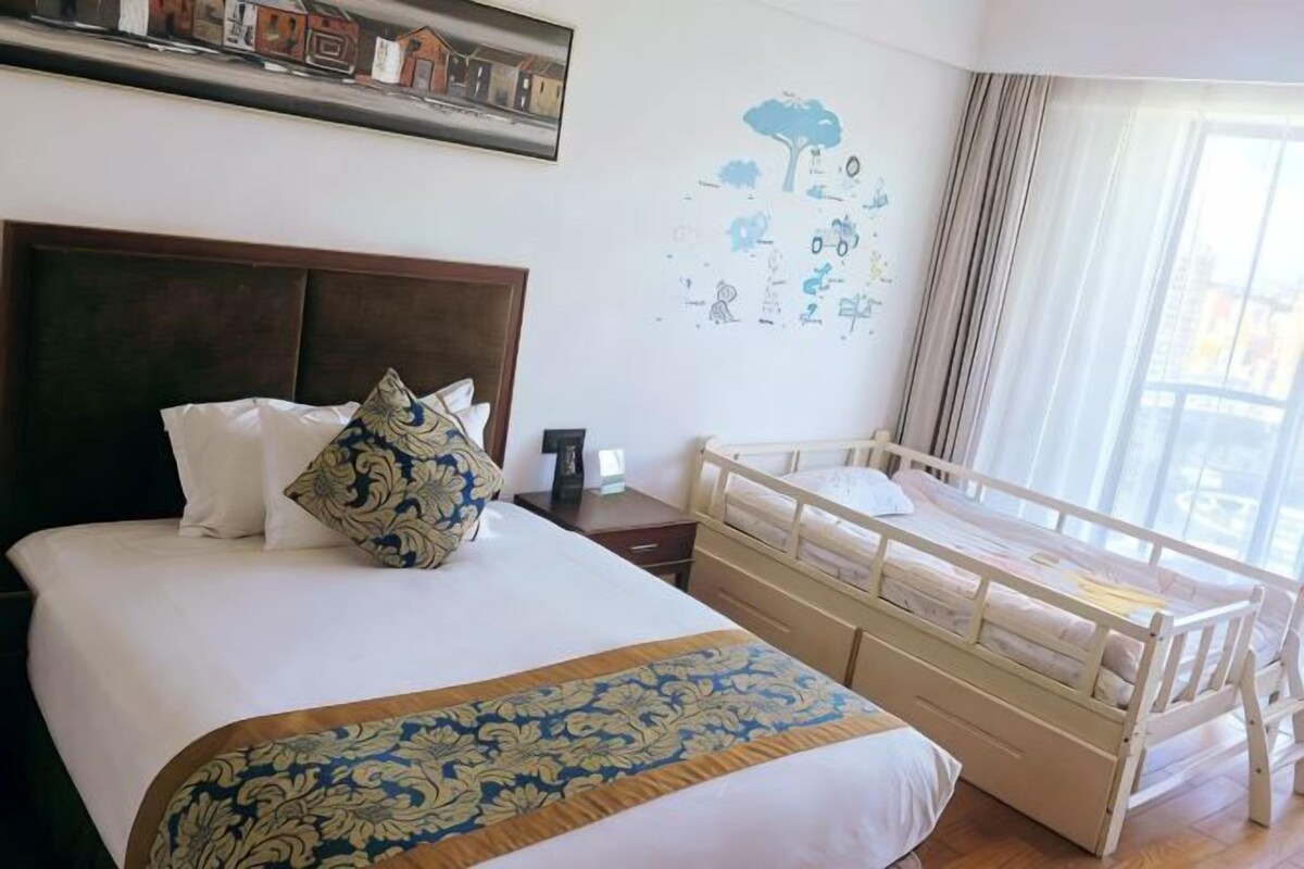 A hotel room with a large bed, a wooden headboard, a small bedside table, a nightstand, a crib, and a window with curtains.