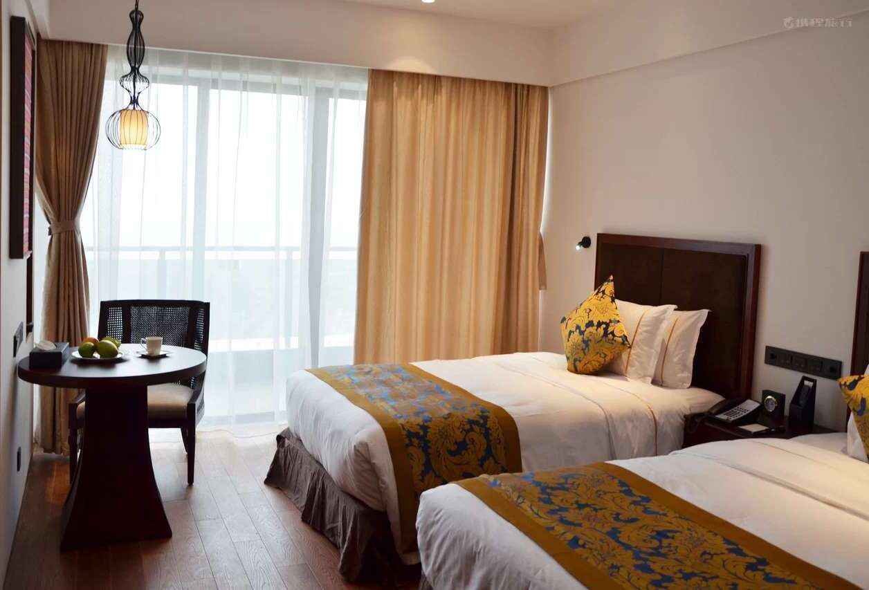 A hotel room with two beds, a small table with a chair, a desk with a telephone, and a large window with curtains.