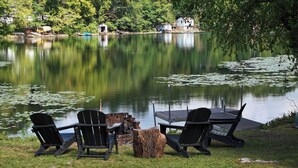 A lakeside seating area with Adirondack chairs and a table, surrounded by a wooden deck and a fire pit. - Lakefront/Legoland/West Point Retreat Under 1 Hour From NYC! Heated pool & Spa! (Monroe)