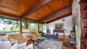 A spacious living area with a wooden ceiling, a stone fireplace, and a large window offering a view of trees.