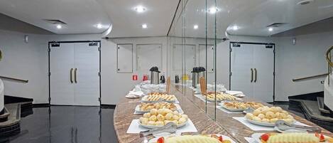 A buffet setup with various food items and utensils on a long counter, with doors and a fire extinguisher in the background.