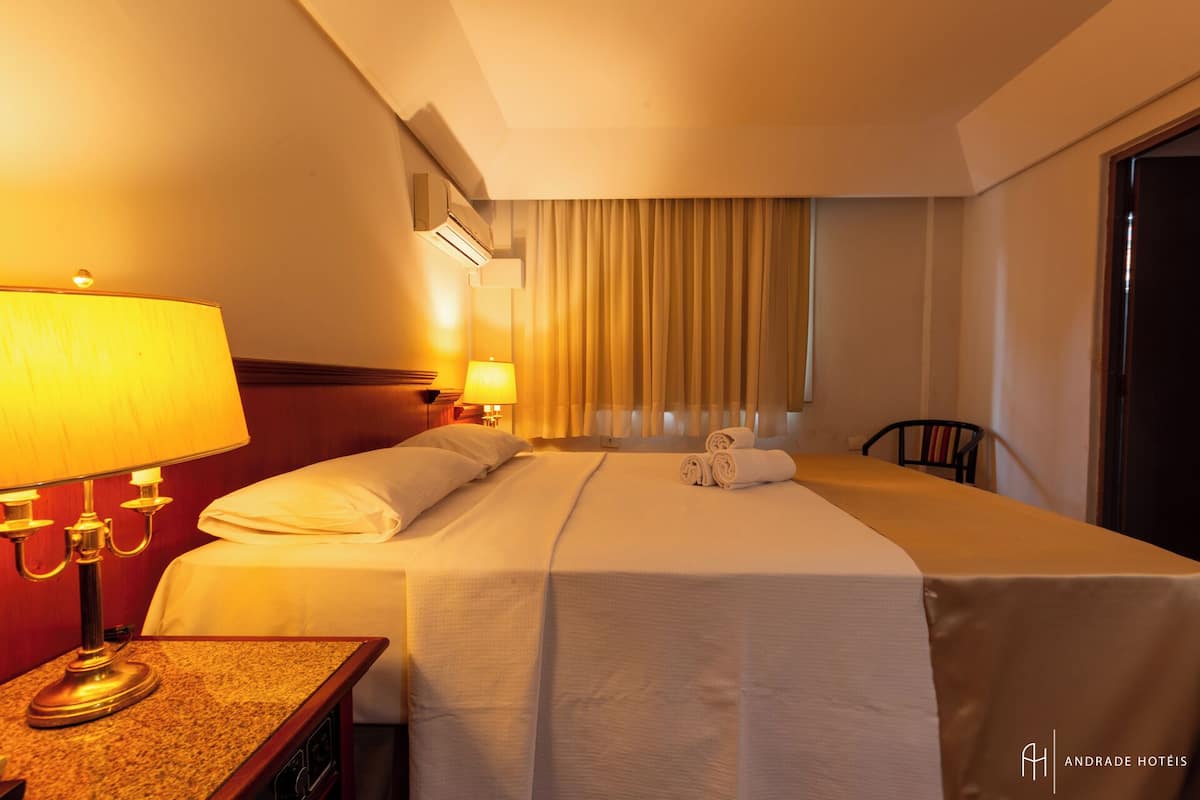 A hotel room with a bed, two bedside lamps, a table with a lamp, a chair, and a window with curtains.