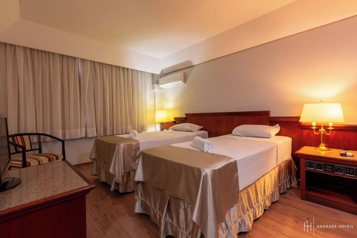 A hotel room with two beds, a desk, a chair, and a nightstand with a lamp.