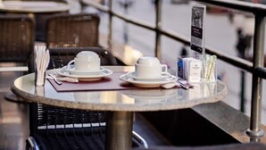 Ein Straßencafé mit Tischen für zwei Personen, bestehend aus weißen Tassen und Untertassen, einem Menüständer und Korbstühlen.