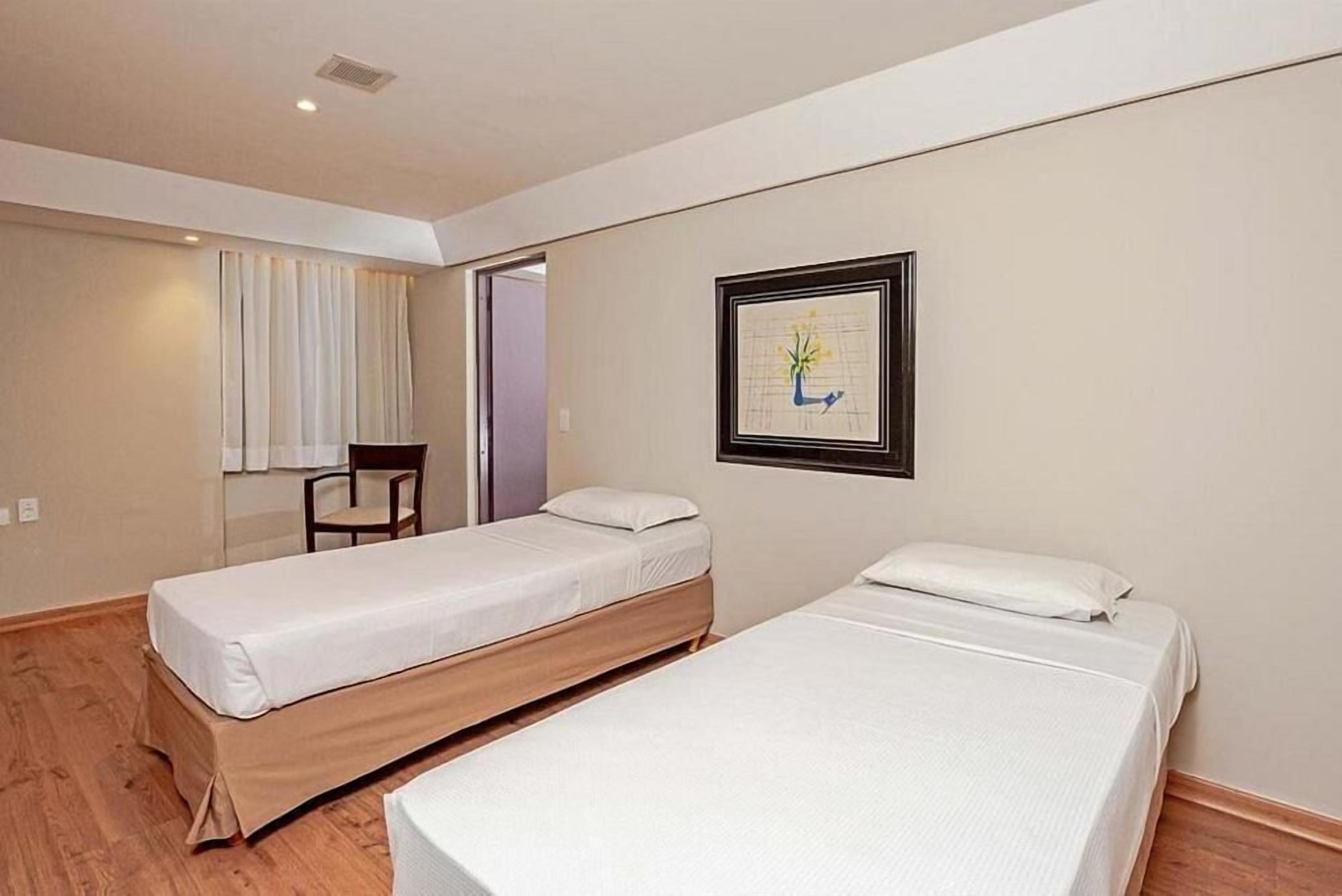 Une chambre avec deux lits simples, une chaise, un tableau encadré au mur et une fenêtre avec des rideaux.