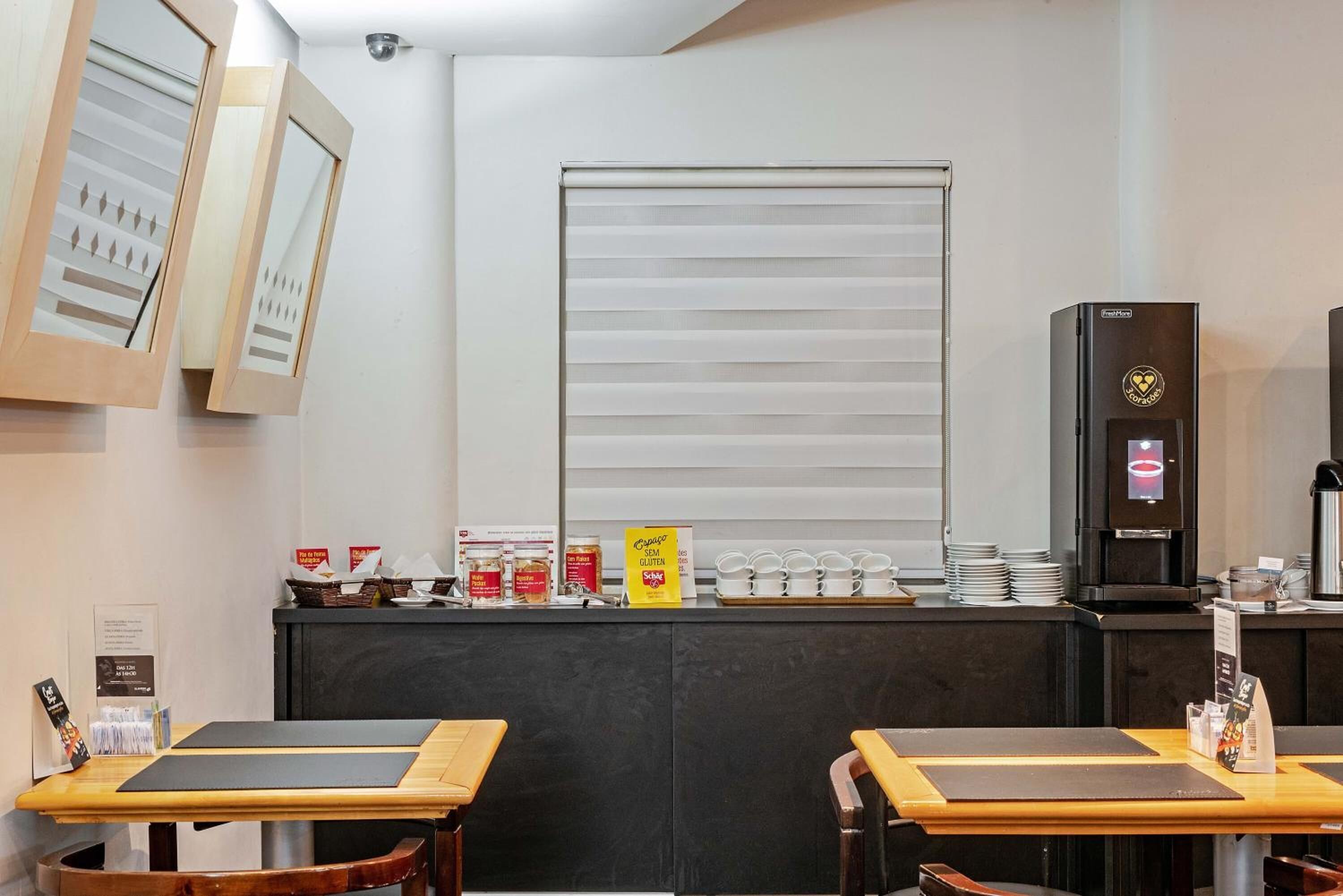 A restaurant with two wooden tables, a counter with a coffee machine, and a window with blinds.