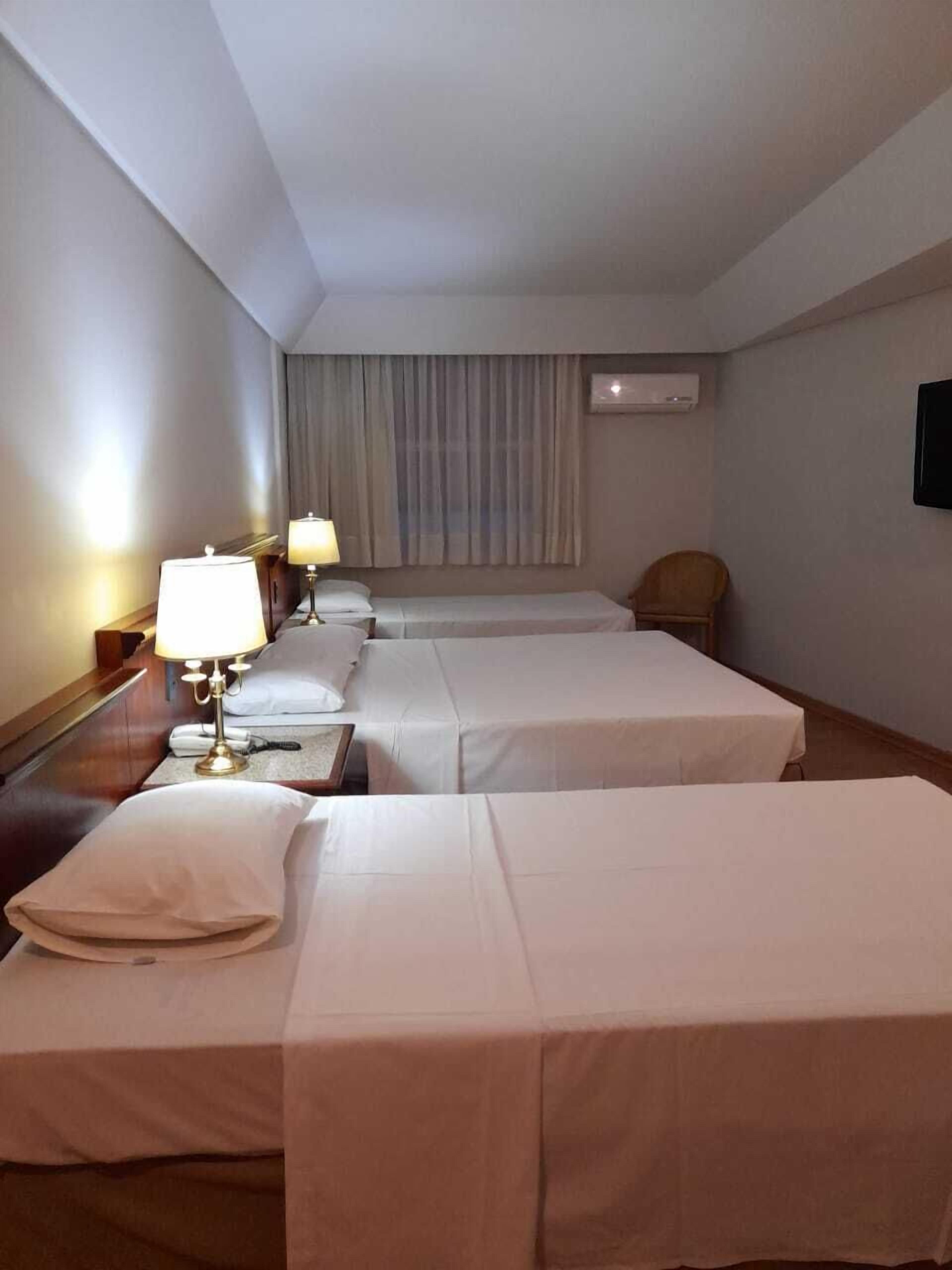 A hotel room with three single beds, a desk with a lamp, a chair, and a TV on the wall.