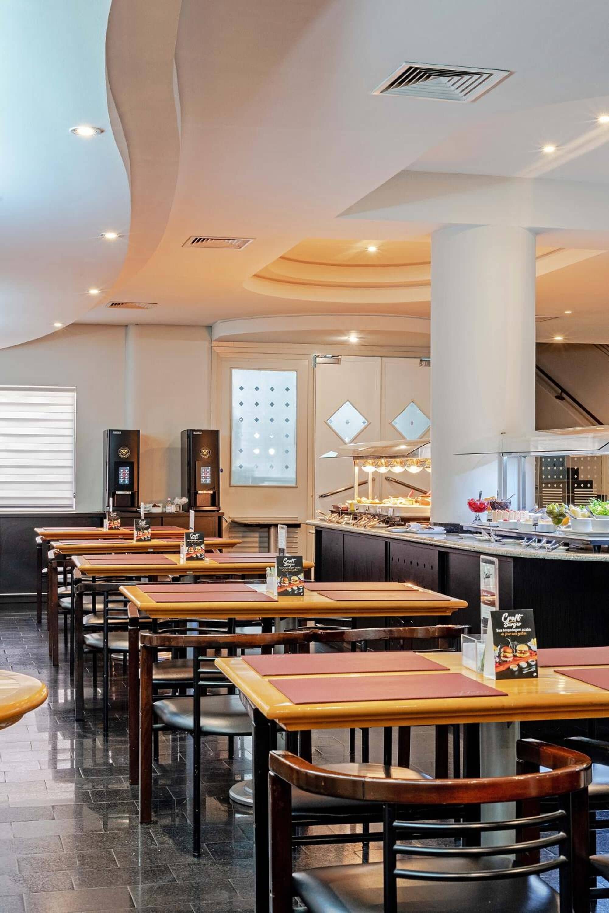 A restaurant with multiple tables and chairs, a buffet counter, and a modern ceiling design.