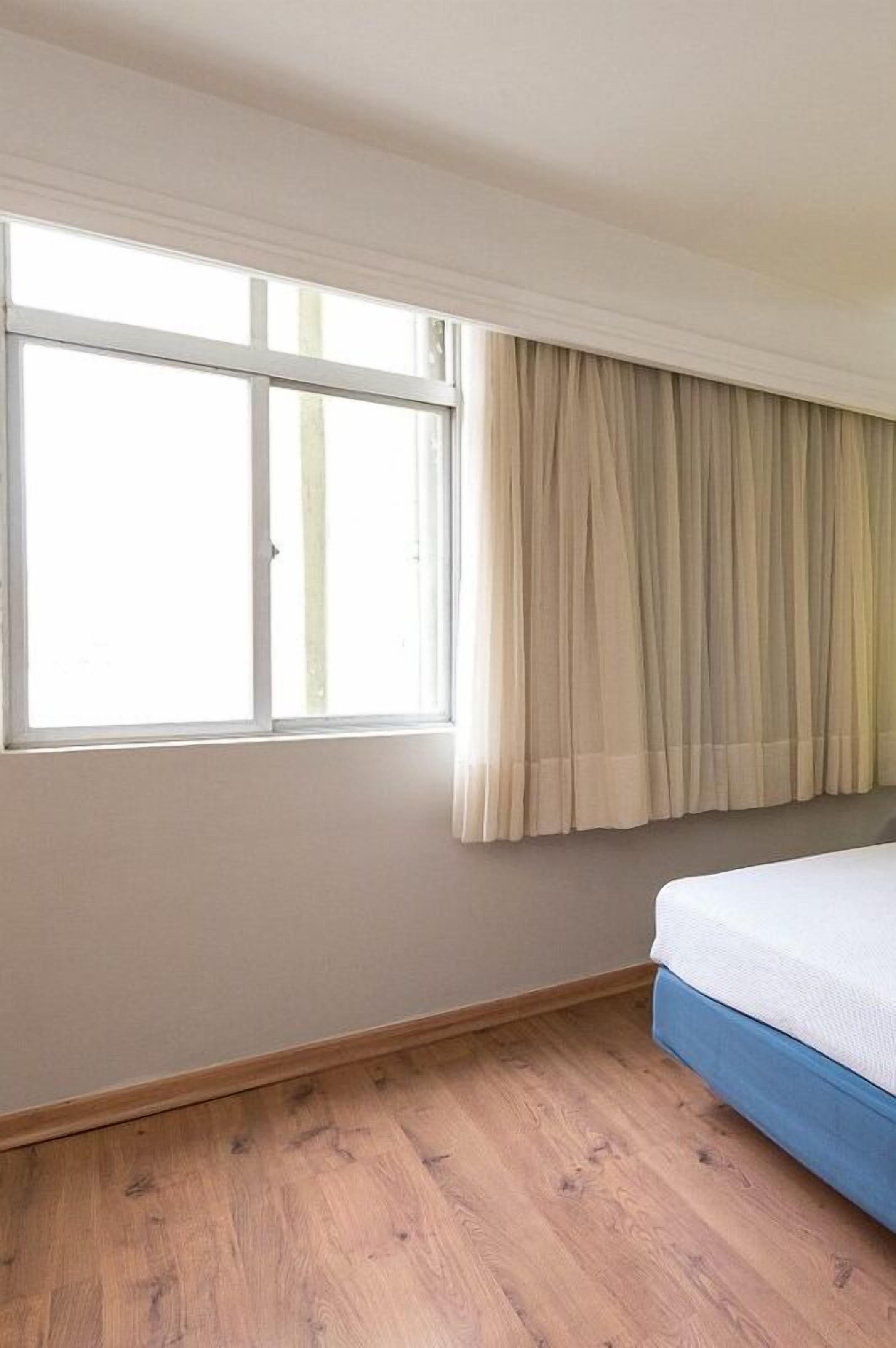 A room with a large window, a bed with a blue headboard, and wooden flooring.
