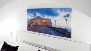 Fotografía de un edificio histórico con un muro fortificado y un camino empedrado que lleva al mar, expuesta en una pared blanca.