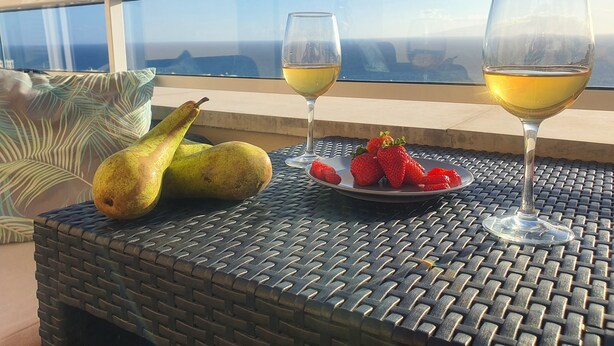 Ett bord med en vävd duk, två glas vitt vin, en tallrik med jordgubbar och två päron på en balkong med fri utsikt över havet.